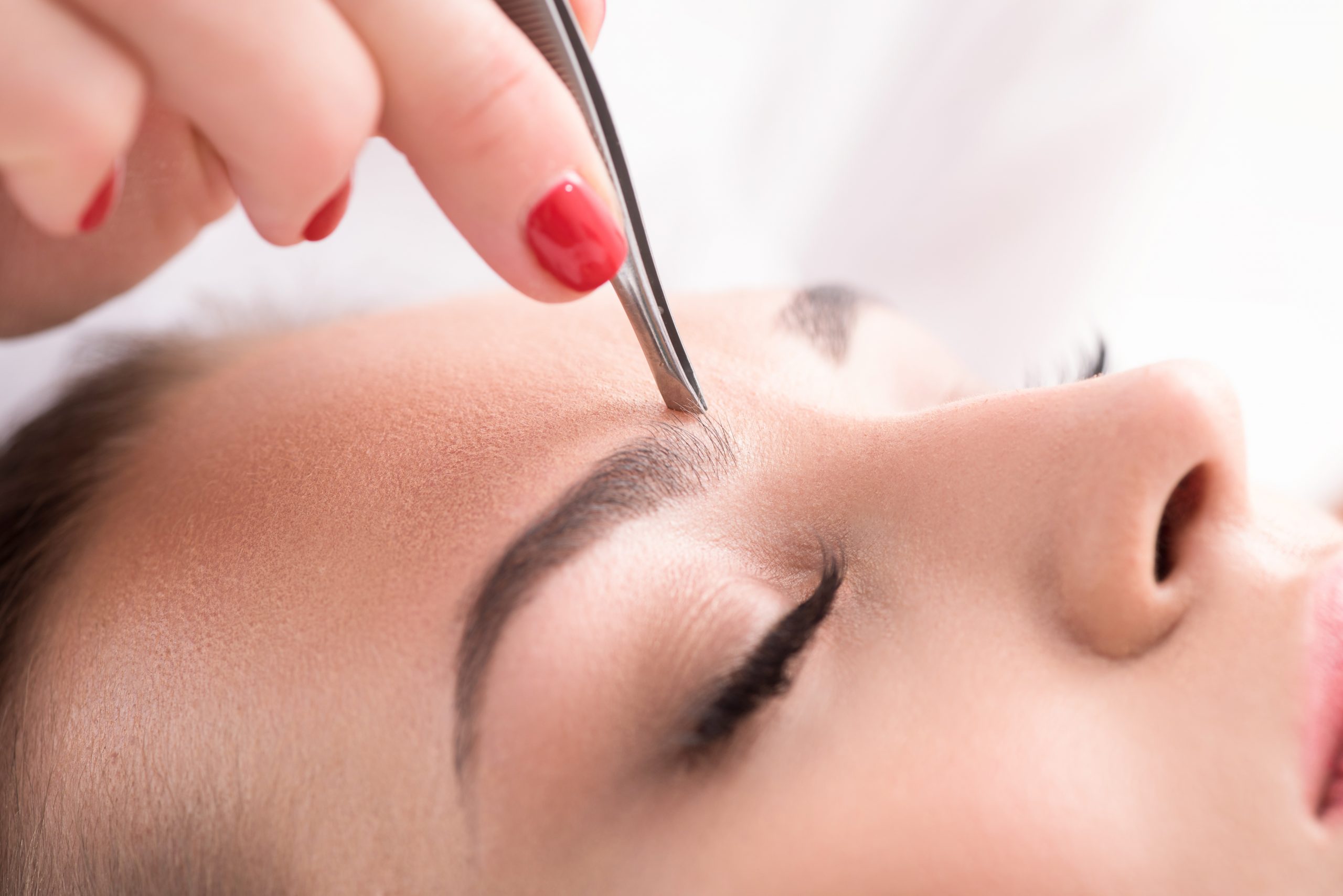 Close up of beautician hand plucking female eyebrow by tweezers