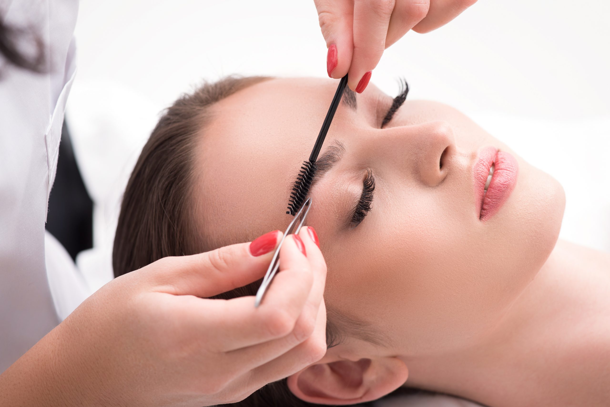 Professional beautician is tweezing female eyebrows and combing it by brush. Young girl is lying with serenity
