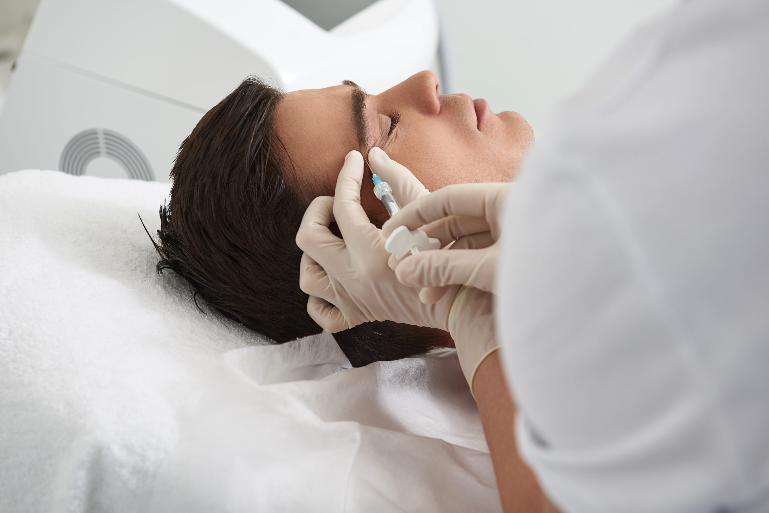 Anti-aging treatment concept. Close up side on portrait of young brunette male having beauty injections procedure of eyebrows zone in cosmetic center