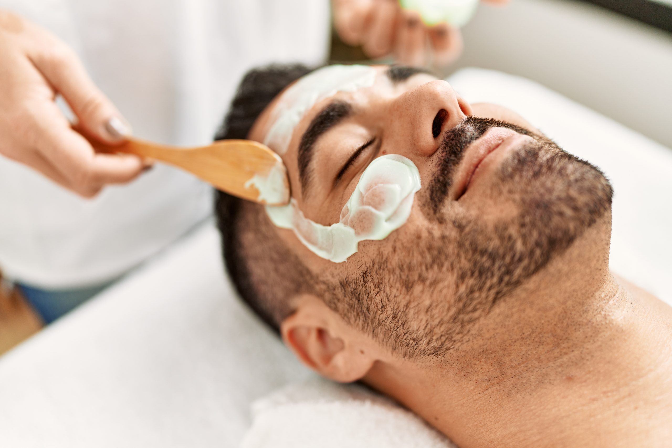 Young hispanic man having facial mask treatment at beauty center