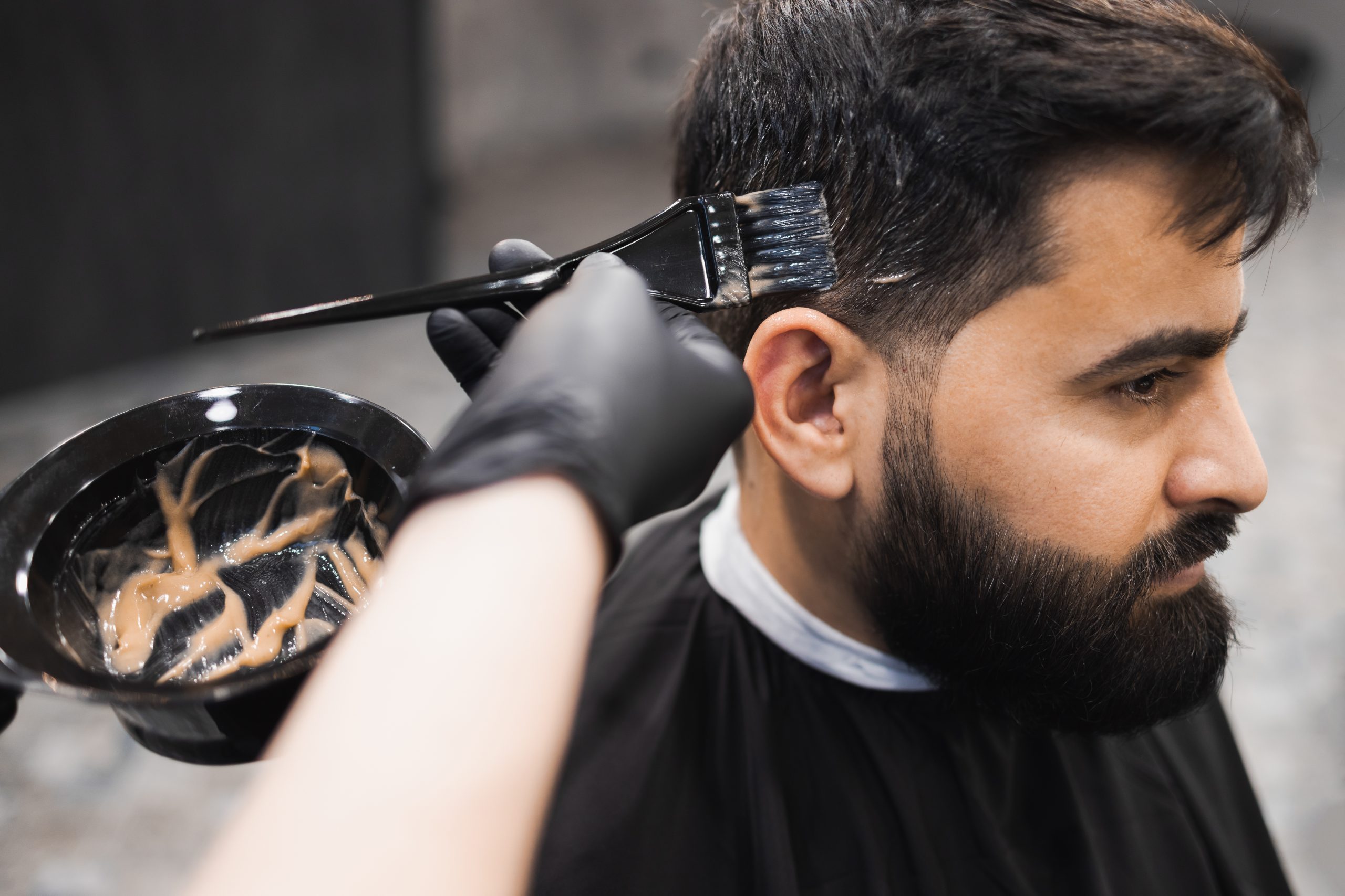 Professional barber dying male client hair in barbershop. Hairdresser applies dye to the client's hair with a brush